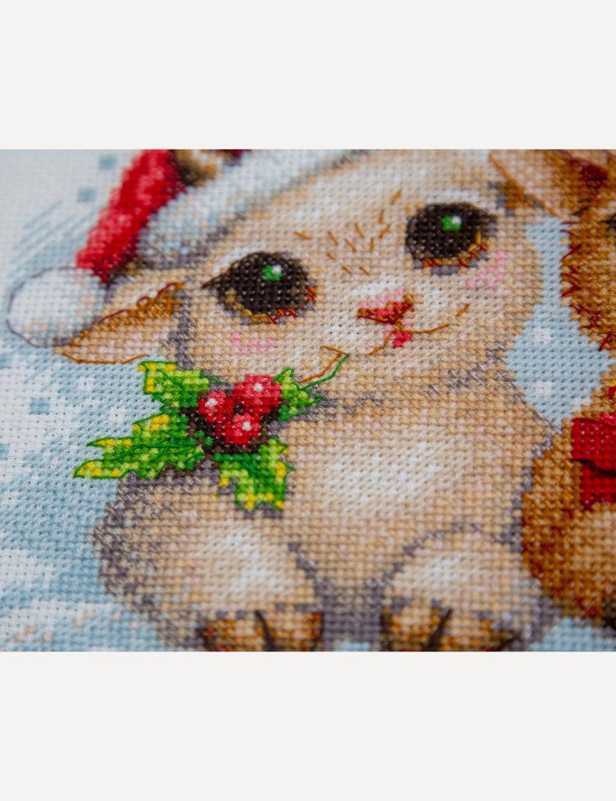 A completed cross-stitch picture of three baby goats wearing red Santa hats, huddled together in falling snow: one with a sprig of holly and red berries, one holding a wrapped Christmas present with a red bow, and one wearing a red-and-green plaid scarf and holding a candy cane.