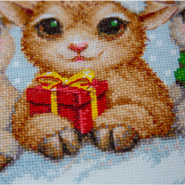A completed cross-stitch picture of three baby goats wearing red Santa hats, huddled together in falling snow: one with a sprig of holly and red berries, one holding a wrapped Christmas present with a red bow, and one wearing a red-and-green plaid scarf and holding a candy cane.
