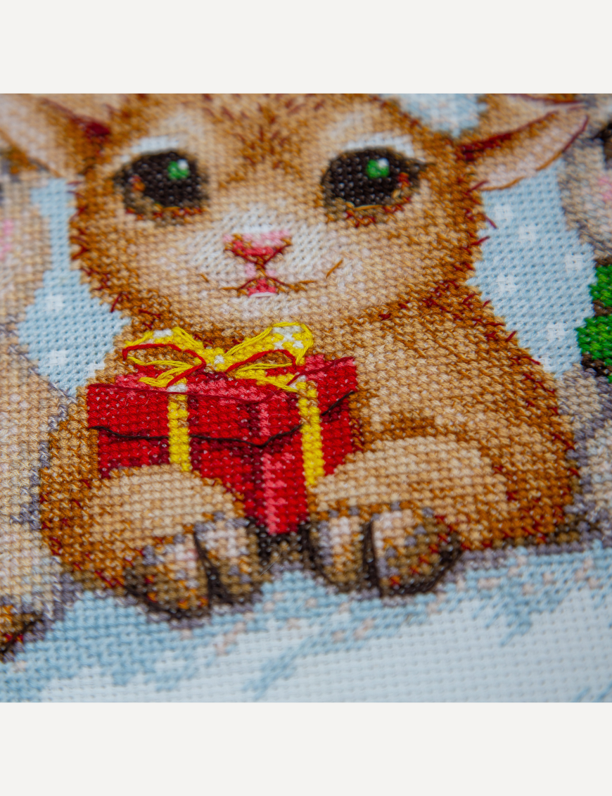 A completed cross-stitch picture of three baby goats wearing red Santa hats, huddled together in falling snow: one with a sprig of holly and red berries, one holding a wrapped Christmas present with a red bow, and one wearing a red-and-green plaid scarf and holding a candy cane.