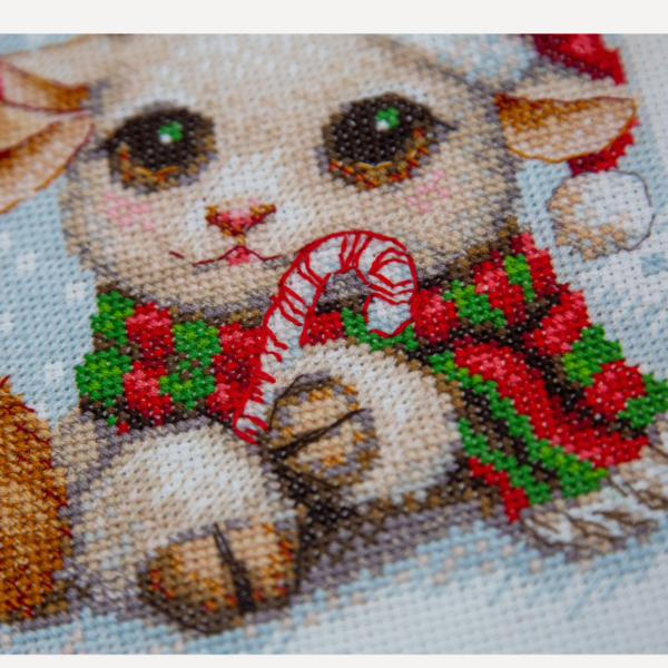 A completed cross-stitch picture of three baby goats wearing red Santa hats, huddled together in falling snow: one with a sprig of holly and red berries, one holding a wrapped Christmas present with a red bow, and one wearing a red-and-green plaid scarf and holding a candy cane.