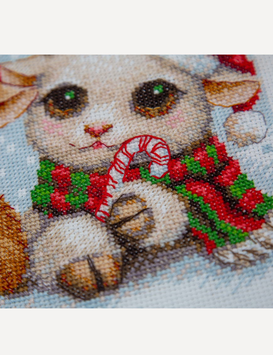 A completed cross-stitch picture of three baby goats wearing red Santa hats, huddled together in falling snow: one with a sprig of holly and red berries, one holding a wrapped Christmas present with a red bow, and one wearing a red-and-green plaid scarf and holding a candy cane.