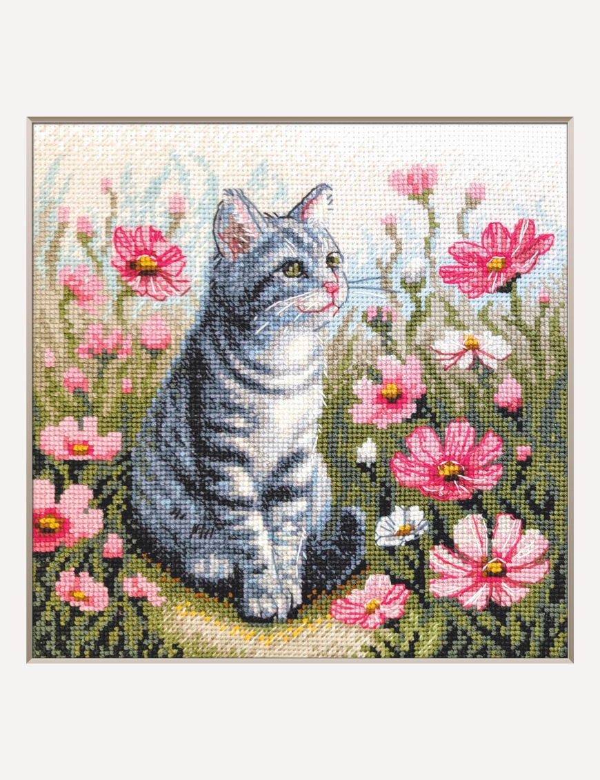 A completed cross-stitch portrait of a grey tabby cat with white markings and green eyes, sitting among tall pink cosmos flowers and a few white blooms, with green foliage and a pale blue sky in the background.