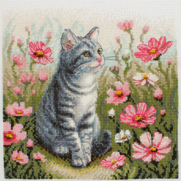 A completed cross-stitch portrait of a grey tabby cat with white markings and green eyes, sitting among tall pink cosmos flowers and a few white blooms, with green foliage and a pale blue sky in the background.