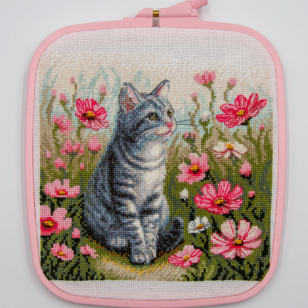 A completed cross-stitch portrait of a grey tabby cat with white markings and green eyes, sitting among tall pink cosmos flowers and a few white blooms, with green foliage and a pale blue sky in the background.