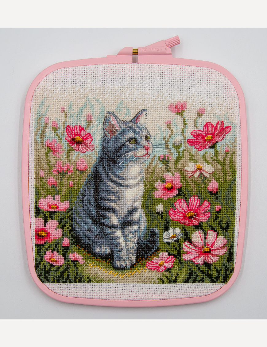 A completed cross-stitch portrait of a grey tabby cat with white markings and green eyes, sitting among tall pink cosmos flowers and a few white blooms, with green foliage and a pale blue sky in the background.