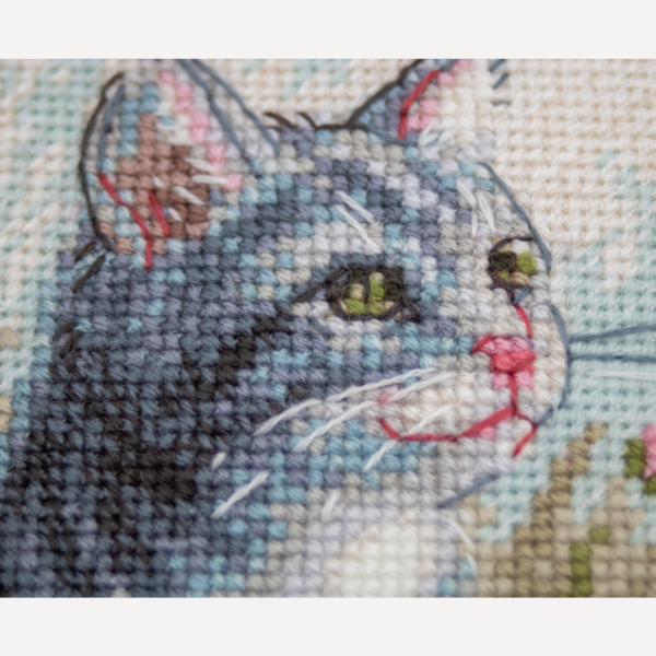 A completed cross-stitch portrait of a grey tabby cat with white markings and green eyes, sitting among tall pink cosmos flowers and a few white blooms, with green foliage and a pale blue sky in the background.