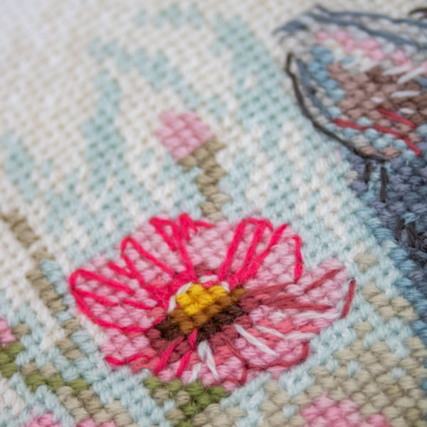 A completed cross-stitch portrait of a grey tabby cat with white markings and green eyes, sitting among tall pink cosmos flowers and a few white blooms, with green foliage and a pale blue sky in the background.