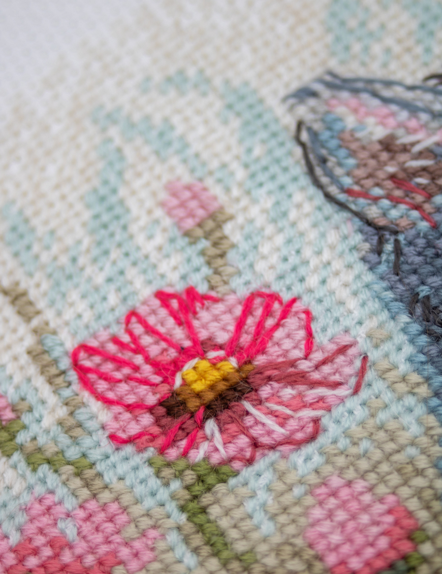 A completed cross-stitch portrait of a grey tabby cat with white markings and green eyes, sitting among tall pink cosmos flowers and a few white blooms, with green foliage and a pale blue sky in the background.