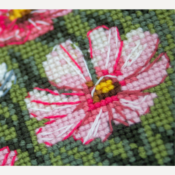 A completed cross-stitch portrait of a grey tabby cat with white markings and green eyes, sitting among tall pink cosmos flowers and a few white blooms, with green foliage and a pale blue sky in the background.