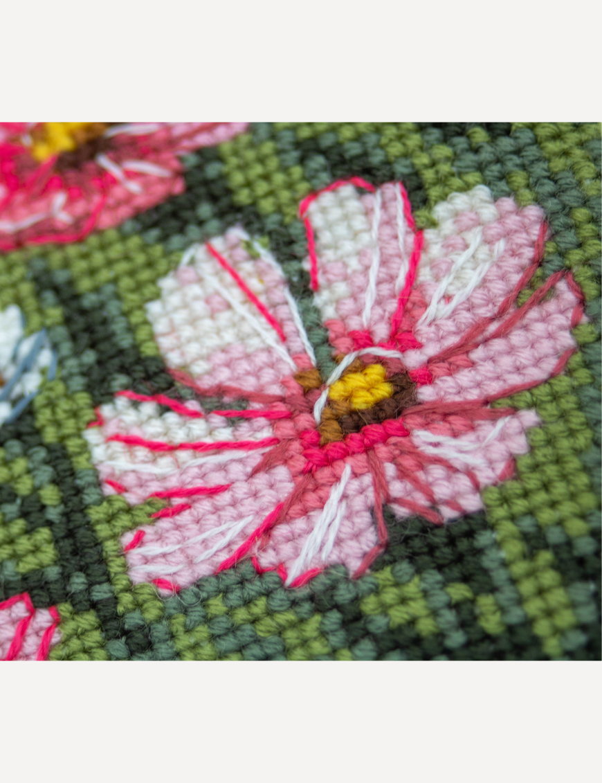 A completed cross-stitch portrait of a grey tabby cat with white markings and green eyes, sitting among tall pink cosmos flowers and a few white blooms, with green foliage and a pale blue sky in the background.