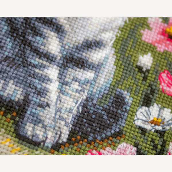 A completed cross-stitch portrait of a grey tabby cat with white markings and green eyes, sitting among tall pink cosmos flowers and a few white blooms, with green foliage and a pale blue sky in the background.