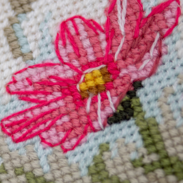 A completed cross-stitch portrait of a grey tabby cat with white markings and green eyes, sitting among tall pink cosmos flowers and a few white blooms, with green foliage and a pale blue sky in the background.