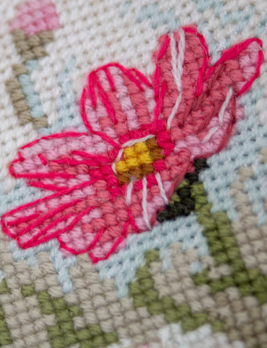 A completed cross-stitch portrait of a grey tabby cat with white markings and green eyes, sitting among tall pink cosmos flowers and a few white blooms, with green foliage and a pale blue sky in the background.