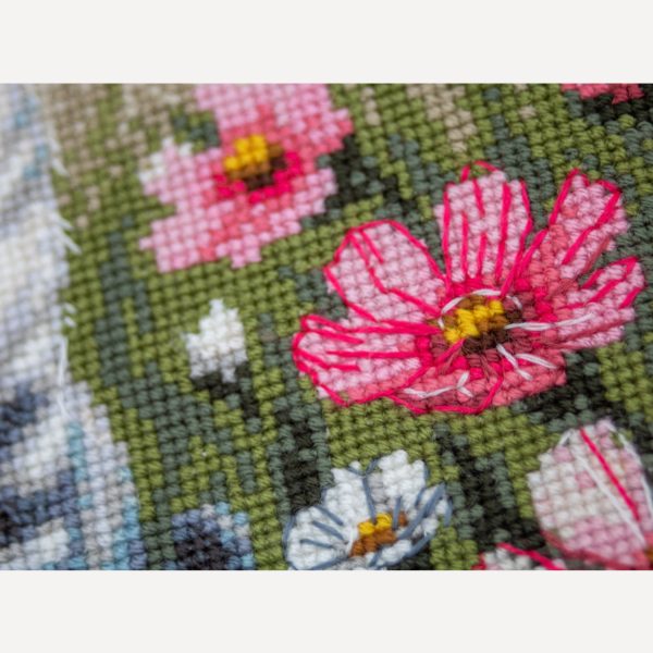 A completed cross-stitch portrait of a grey tabby cat with white markings and green eyes, sitting among tall pink cosmos flowers and a few white blooms, with green foliage and a pale blue sky in the background.