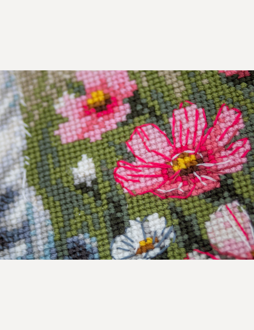 A completed cross-stitch portrait of a grey tabby cat with white markings and green eyes, sitting among tall pink cosmos flowers and a few white blooms, with green foliage and a pale blue sky in the background.