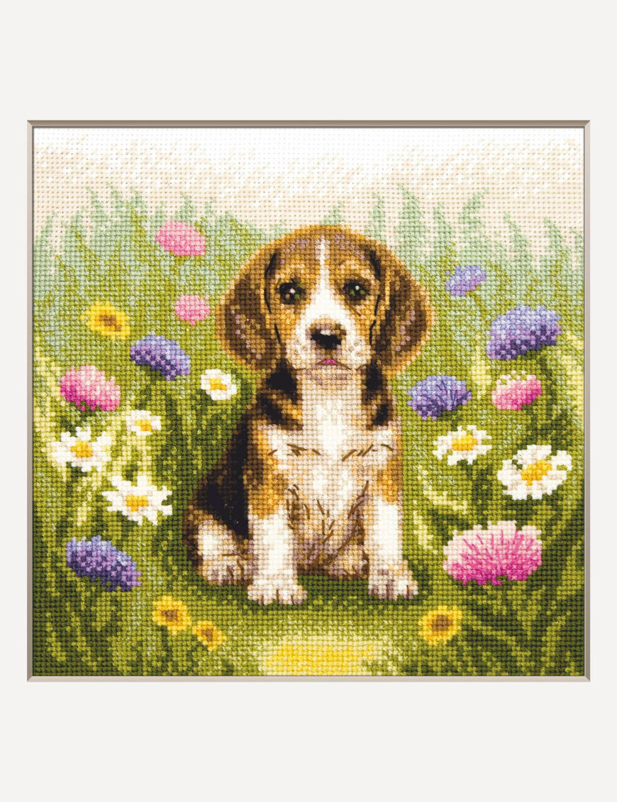A completed cross-stitch portrait of a tricolor beagle puppy with long floppy ears sitting in a green meadow, surrounded by pink and purple wildflowers, white daisies and yellow blooms.