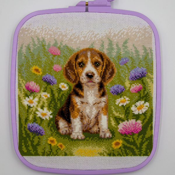 A completed cross-stitch portrait of a tricolor beagle puppy with long floppy ears sitting in a green meadow, surrounded by pink and purple wildflowers, white daisies and yellow blooms.