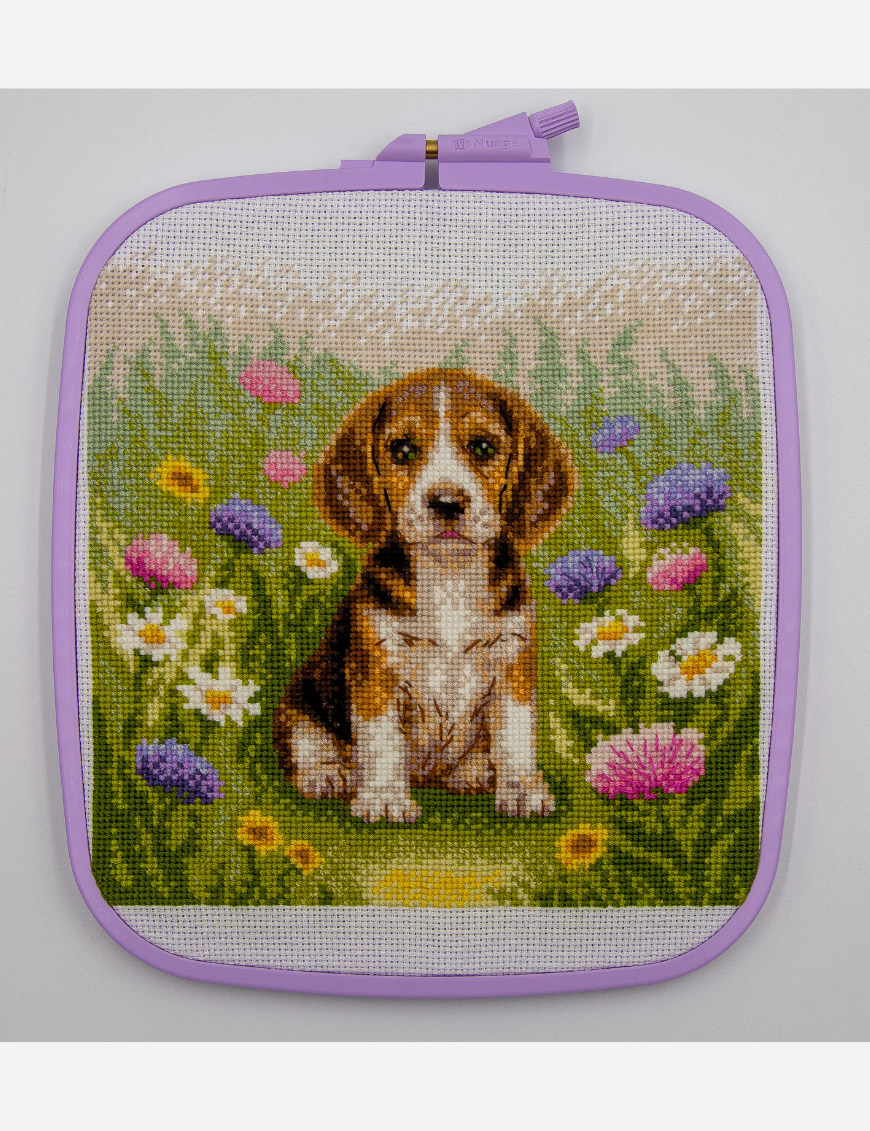 A completed cross-stitch portrait of a tricolor beagle puppy with long floppy ears sitting in a green meadow, surrounded by pink and purple wildflowers, white daisies and yellow blooms.
