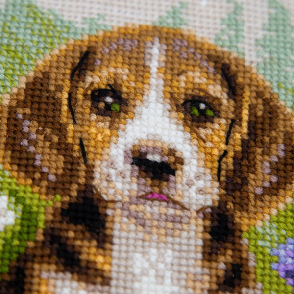 A completed cross-stitch portrait of a tricolor beagle puppy with long floppy ears sitting in a green meadow, surrounded by pink and purple wildflowers, white daisies and yellow blooms.
