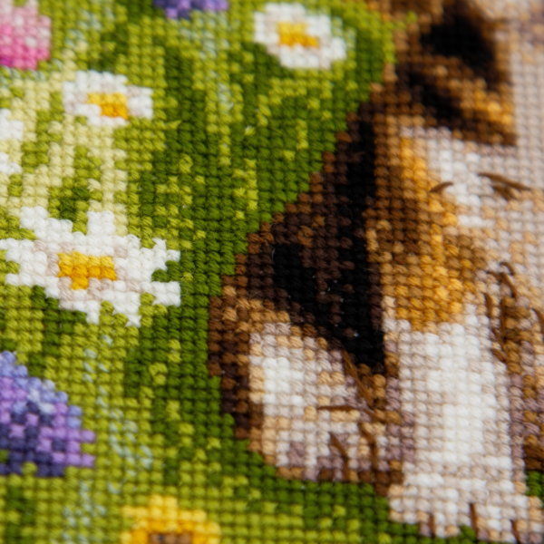 A completed cross-stitch portrait of a tricolor beagle puppy with long floppy ears sitting in a green meadow, surrounded by pink and purple wildflowers, white daisies and yellow blooms.