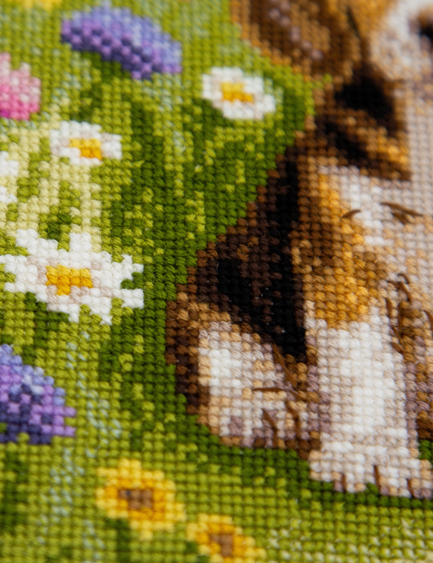 A completed cross-stitch portrait of a tricolor beagle puppy with long floppy ears sitting in a green meadow, surrounded by pink and purple wildflowers, white daisies and yellow blooms.