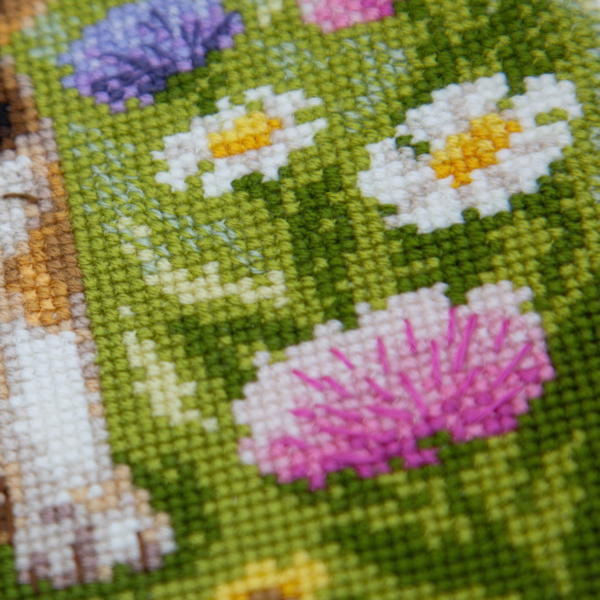 A completed cross-stitch portrait of a tricolor beagle puppy with long floppy ears sitting in a green meadow, surrounded by pink and purple wildflowers, white daisies and yellow blooms.