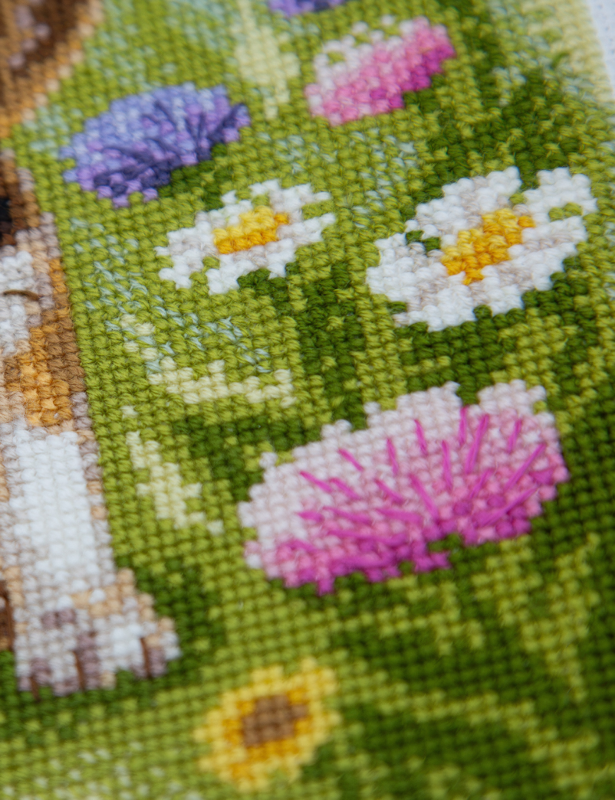 A completed cross-stitch portrait of a tricolor beagle puppy with long floppy ears sitting in a green meadow, surrounded by pink and purple wildflowers, white daisies and yellow blooms.