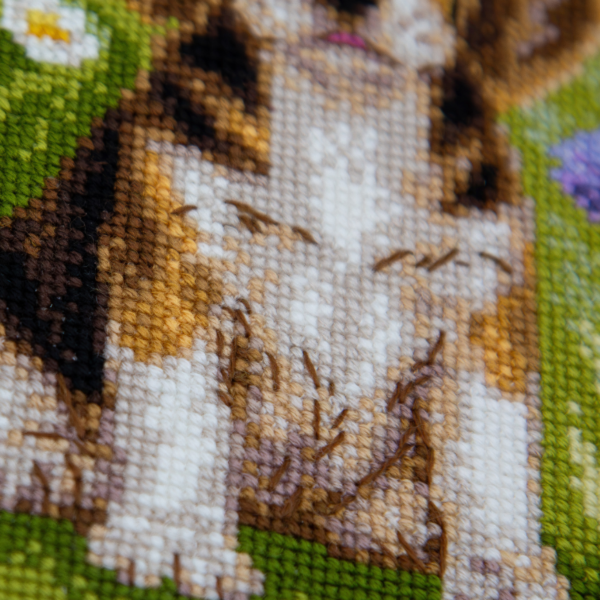 A completed cross-stitch portrait of a tricolor beagle puppy with long floppy ears sitting in a green meadow, surrounded by pink and purple wildflowers, white daisies and yellow blooms.
