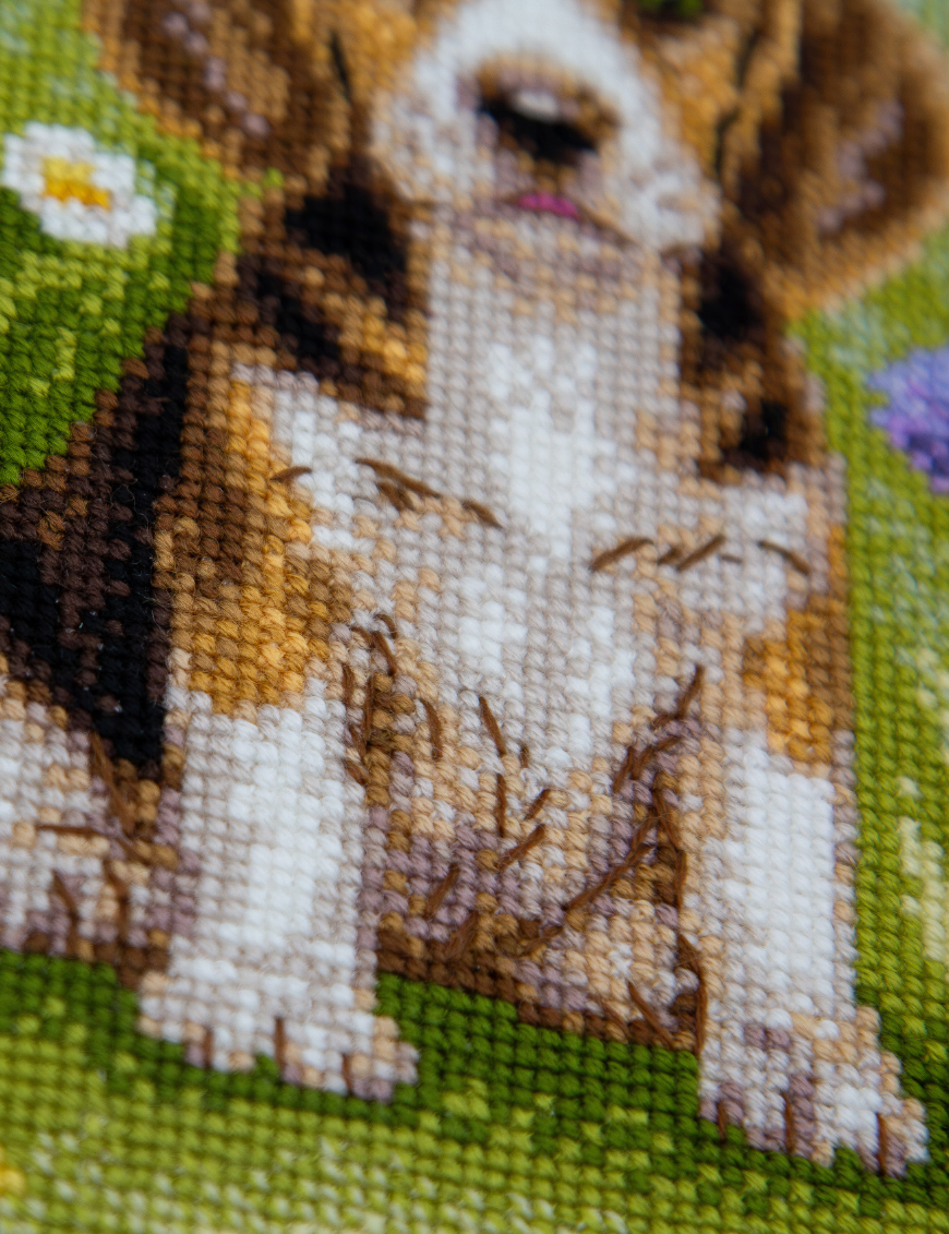 A completed cross-stitch portrait of a tricolor beagle puppy with long floppy ears sitting in a green meadow, surrounded by pink and purple wildflowers, white daisies and yellow blooms.