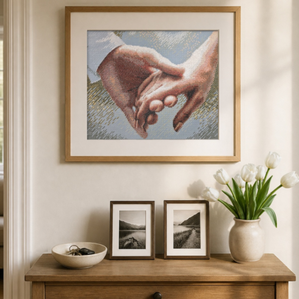 The finished cross-stitch of two clasped hands in a large oak frame above a wooden console table with white tulips in a vase and two small framed black-and-white photographs.