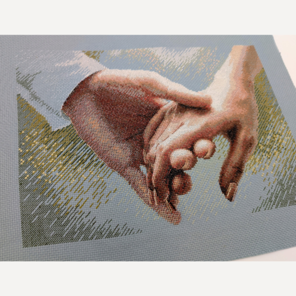 The finished cross-stitch of two clasped hands on blue Aida fabric, photographed at an angle without a frame, showing the shaded skin tones and golden sparkles in the background.