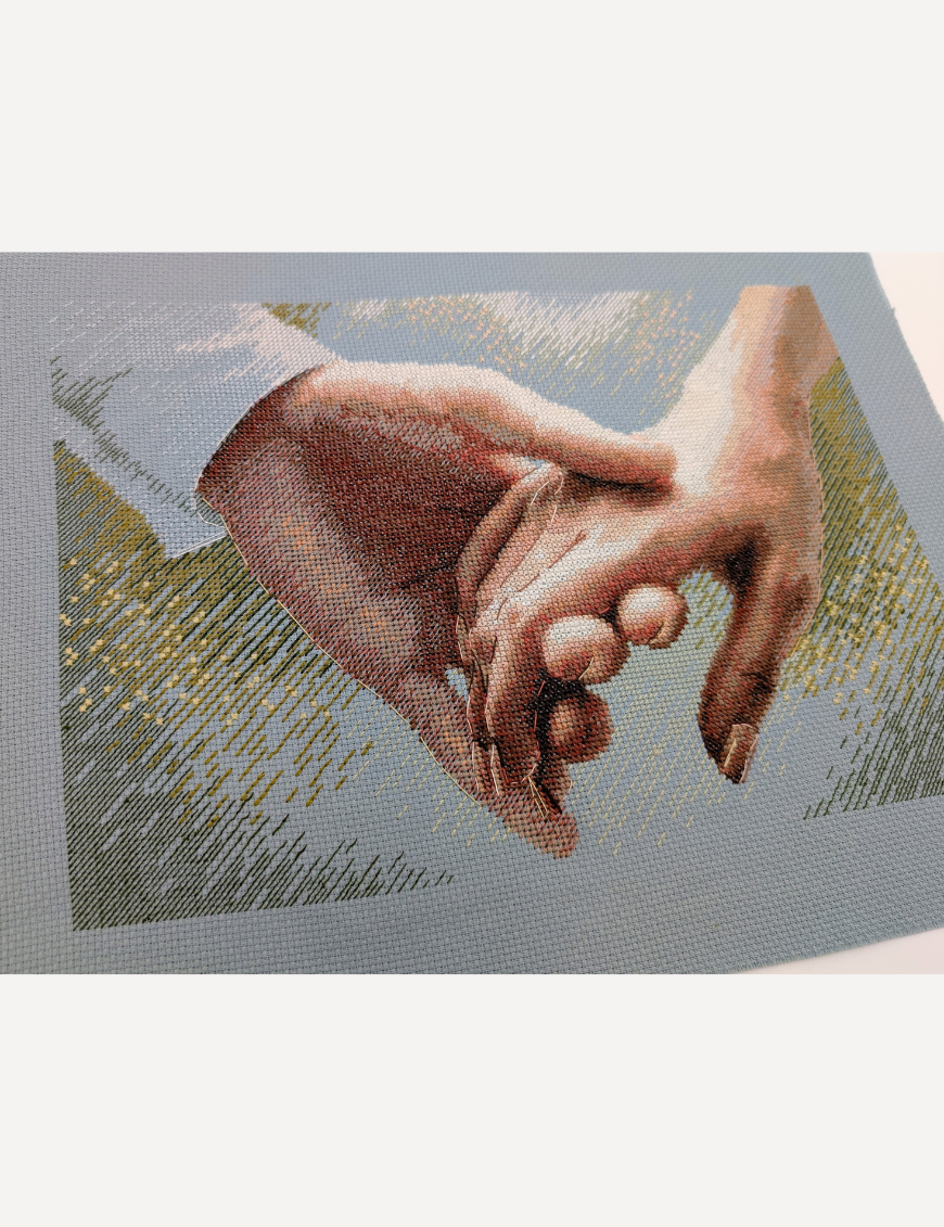 The finished cross-stitch of two clasped hands on blue Aida fabric, photographed at an angle without a frame, showing the shaded skin tones and golden sparkles in the background.