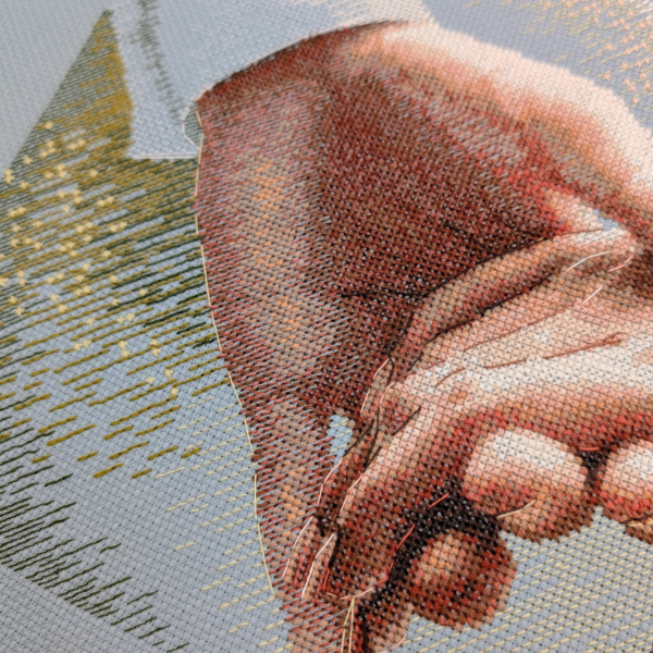 Angled close-up of the cross-stitched hand and white shirt cuff on blue Aida, with golden sparkle stitches and green shading in the background.