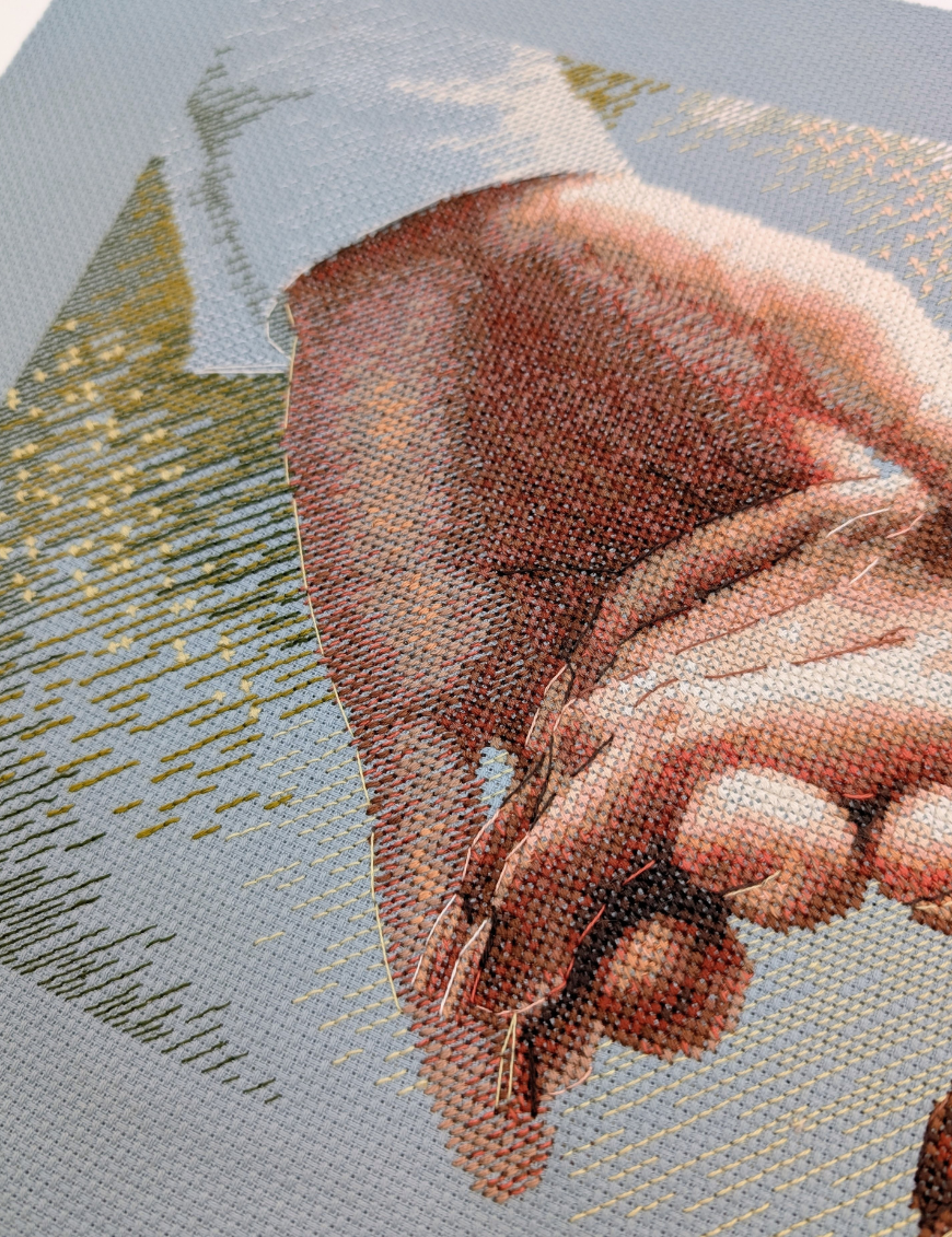 Angled close-up of the cross-stitched hand and white shirt cuff on blue Aida, with golden sparkle stitches and green shading in the background.