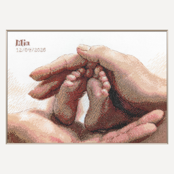 A completed cross-stitch picture of a newborn baby’s tiny feet cradled between a parent’s hands in warm terracotta, peach and rose tones, with space at the top left for a name and date.