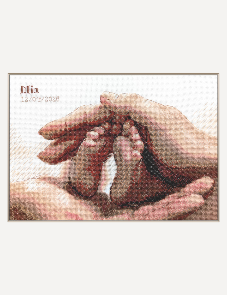 A completed cross-stitch picture of a newborn baby’s tiny feet cradled between a parent’s hands in warm terracotta, peach and rose tones, with space at the top left for a name and date.