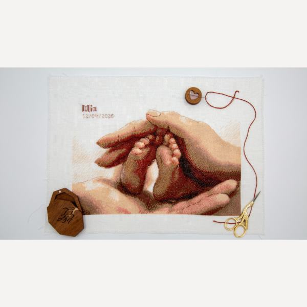 A completed cross-stitch picture of a newborn baby’s tiny feet cradled between a parent’s hands in warm terracotta, peach and rose tones, with space at the top left for a name and date. Shown unframed on Aida fabric with a wooden needle minder, red thread and gold stork scissors.