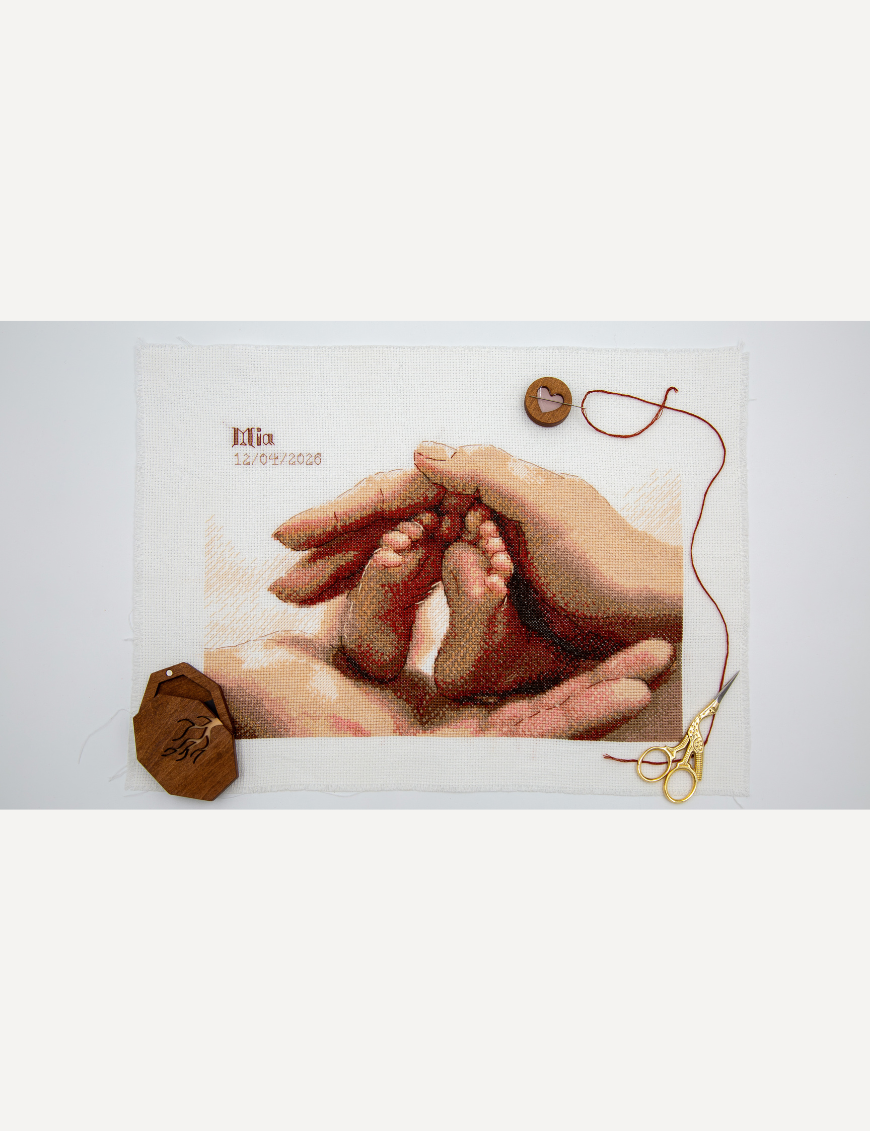 A completed cross-stitch picture of a newborn baby’s tiny feet cradled between a parent’s hands in warm terracotta, peach and rose tones, with space at the top left for a name and date. Shown unframed on Aida fabric with a wooden needle minder, red thread and gold stork scissors.