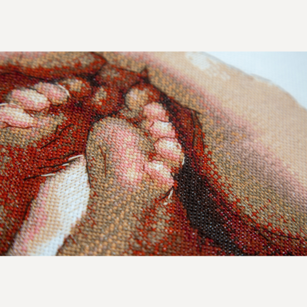 Angled close-up of a cross-stitched baby foot and toes cradled in a hand, in warm red, peach and cream threads.