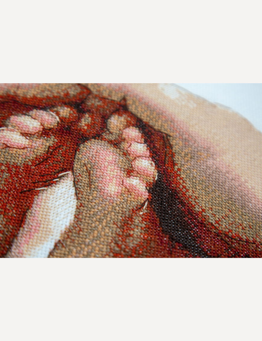 Angled close-up of a cross-stitched baby foot and toes cradled in a hand, in warm red, peach and cream threads.